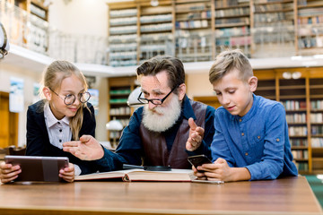 Little school-age smiling children, boy and girl, comparing tablet e-reader and smartphone with their grandfathers traditional book. Elderly bearded man is trying to convince kids to read a book