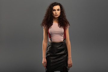 Young beautiful girl with curls on her head with a serious facial expression on a gray background. Portrait of a woman with a strong face with brown eyes in a pink T-shirt and red suspenders.