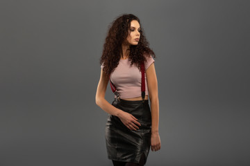 Young beautiful girl with curls on her head with a serious facial expression on a gray background. Portrait of a woman with a strong face with brown eyes in a pink T-shirt and red suspenders.