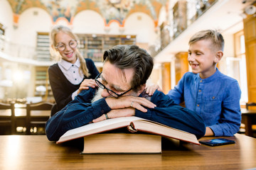 Funny laughing children, boy and girl, waking up elderly man librarian or grandfather sitting at the table and sleeping on the books. Vintage library interior