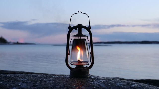 Old kerosene lamp on the rock. Vintage lantern lighting in dark. Sea and sunset sky on background. Smoke comes from the top.