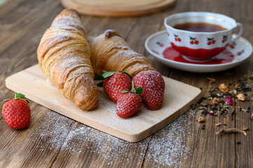 Two croissants on a wooden cutting board with strawberries and a cup of tea