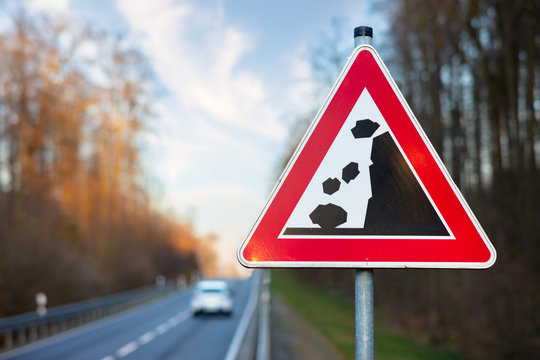 A triangular red and white warning sign on a country road in Germany. The sign warns of falling rocks and slipping soil. In the background out of focus with bokeh a moving car.