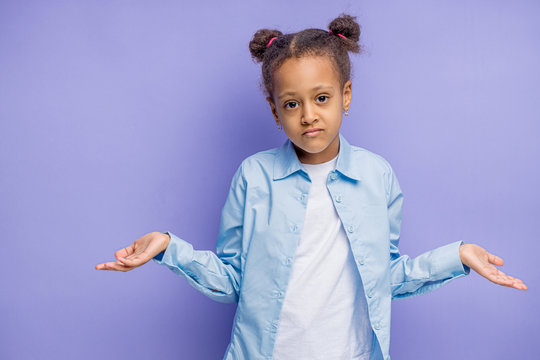 Portrait Of Hesitant African Mulatto Child, She Looks Clueless, Being Puzzled, Shrugs Shoulders, Isolated Over Purple Background. Doubts And Puzzlement Concept