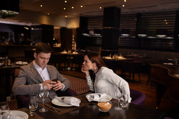 indignant lady and offended male sit in restaurant. caucasian couple spend their evening together, but they are unhappy, have quarrel. male look at screen of mobile phone