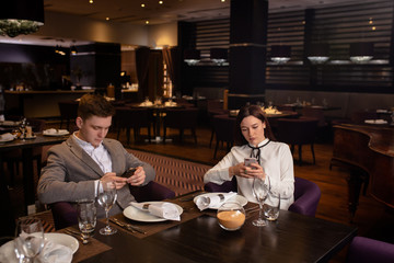 bad evening in a restaurant. young caucasian couple quarreled in representative luxury restaurant, they don't talk with each other, sit looking at their own mobile phone