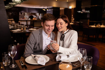 young caucasian couple have dinner together, look at smartphone in restaurant. woman show cool video to boyfriend. indoors. man and woman enjoy spending time together