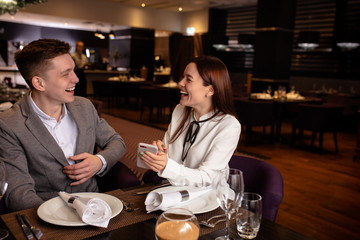 attractive lady happily telling interesting story to her boyfriend while they have dinner in luxury restaurant, they are on date, she show on smartphone and laugh