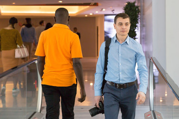 A male photographer is walking inside a building among people. A man with a camera in his hand is among passers-by in shopping business center.