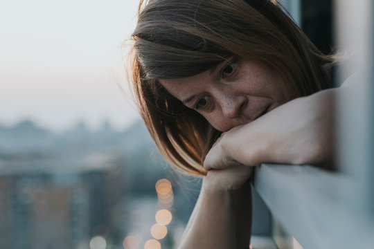 Young Sad Woman Looking Outside Through Balcony Of An Apartment Building