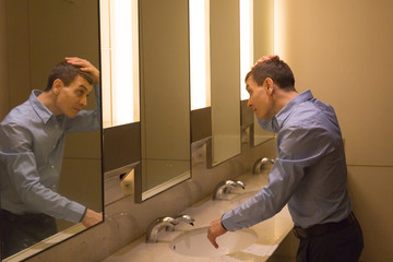An adult Caucasian man in a blue shirt is standing in front of a toilet mirror and combing his head...