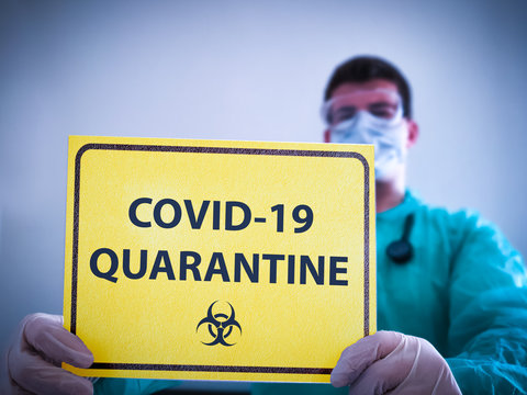 COVID 19 Coronavirus, Doctor In Isolation Holdin Yellow Danger Sign Of Corona Virus Quarantine. Quarantine Alert Sign In Hospital With Blurred Doctor With Mask And Protective Suit. Korona Virus 