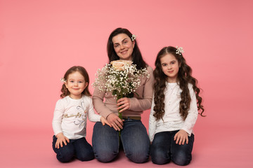 Beautiful family in casual clothes, with white flowers in their hair sitting on the ground, mom holding bouquet of spring flowers and looking at the camera