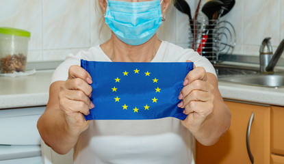 Close-up of a matured woman in a medical mask with an EU flag sitting at home in the kitchen. Quarantine Europe - concept.