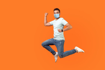 Fototapeta na wymiar Full length of positive inspired brunette man with medical mask in sneakers, denim outfit jumping in air or running quickly fast. indoor studio shot isolated on orange background, empty copy space