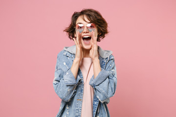 Cheerful young brunette woman girl in casual denim jacket, eyeglasses posing isolated on pastel pink background. People lifestyle concept. Mock up copy space. Screaming with hands gesture near mouth.