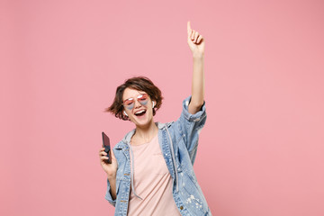 Cheerful young brunette woman in casual denim jacket eyeglasses isolated on pastel pink background. People lifestyle concept. Mock up copy space. Listen music with air pods hold mobile phone, dancing. - obrazy, fototapety, plakaty