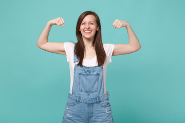 Fototapeta premium Strong smiling young brunette woman girl in casual denim clothes posing isolated on blue turquoise background studio portrait. People lifestyle concept. Mock up copy space. Showing biceps, muscles.