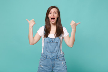 Surprised young brunette woman girl in casual denim clothes posing isolated on blue turquoise wall background studio portrait. People lifestyle concept. Mock up copy space. Pointing thumbs aside.