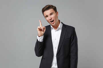 Displeased young business man in classic black suit shirt posing isolated on grey wall background. Achievement career wealth business concept. Mock up copy space. Pointing index finger up, swearing.