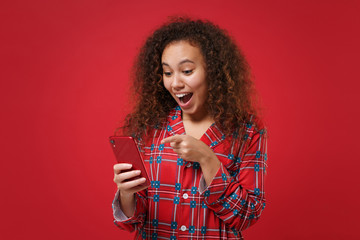 Excited young african american girl in pajamas homewear posing resting at home isolated on red background. Relax good mood lifestyle concept. Mock up copy space. Pointing index finger on mobile phone.