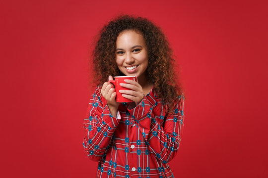 Smiling young african american girl in pajamas homewear posing while resting at home isolated on red wall background. Relax good mood lifestyle concept. Mock up copy space. Hold cup of coffee or tea.