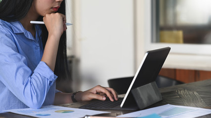 Cropped image of designer woman holding a stylus pen while sitting in front her computer tablet with keyboard case at the modern working table over smart office as background.