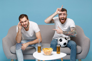 Displeased excited funny men guys friends in white t-shirt sit on couch isolated on blue background. Sport leisure concept. Cheer up support favorite team with ball point aside showing loser gesture.