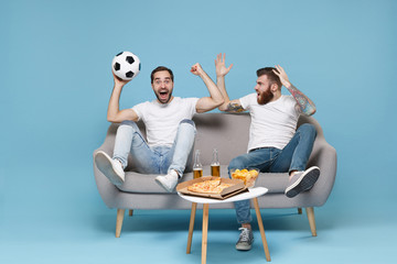 Excited men guys friends in white t-shirt sit on couch isolated on blue background. Sport leisure...