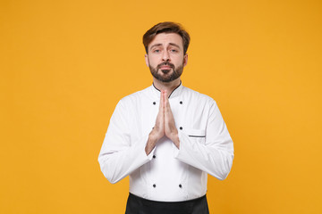 Pleading young bearded male chef cook or baker man in white uniform shirt posing isolated on yellow background studio portrait. Cooking food concept. Mock up copy space. Hold hands folded in prayer.