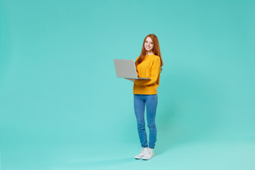 Smiling young redhead girl in yellow knitted sweater posing isolated on blue turquoise background studio portrait. People emotions lifestyle concept. Mock up copy space. Work on laptop pc computer.