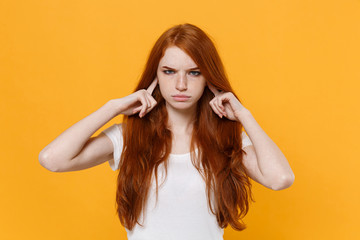 Displeased young redhead woman girl in white blank empty t-shirt posing isolated on yellow wall background studio portrait. People lifestyle concept. Mock up copy space. Covering ears with fingers.