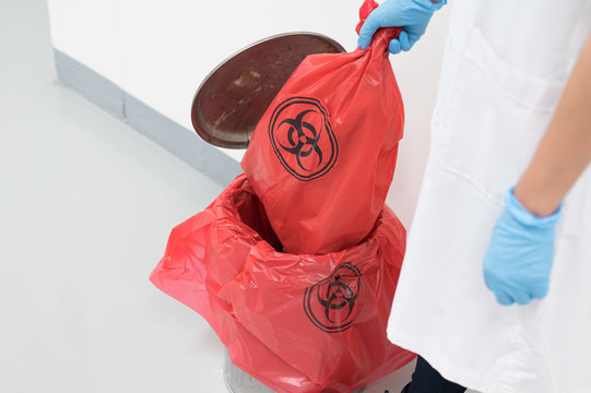 Scientist wearing blue gloves and red bag with bioharzard sign.A woman worker hand holding red garbage bag.Maid and infection waste bin at the indoor public building.Infectious control.