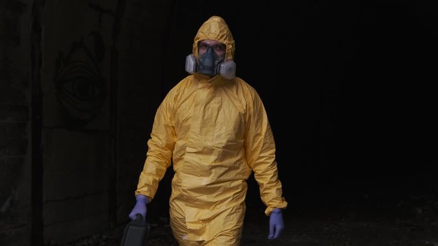 A man in a yellow hazmat suit and protective mask with a suitcase comes out of the tunnel. Tracking Shot.