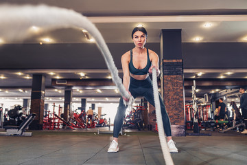 Fototapeta premium Caucasian fit woman dressed in sportsoutfit posing with battle ropes at gym.