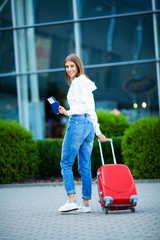 A woman with a passport and a red suitcase near the airport is going on a trip
