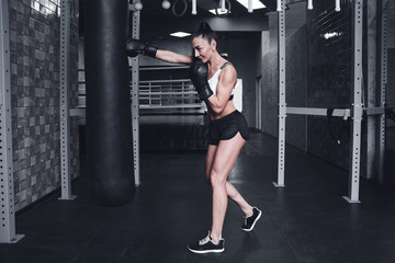 Fototapeta na wymiar Fit sexy strong brunette woman in sportswear top and shorts fights a punching bag with boxing gloves in the gym