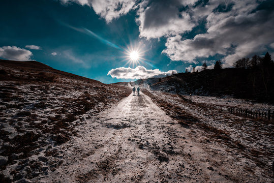 Walking to heaven. In a autumnal landscape, a meadow with dry (brown) grass, a blue sky with clouds and trekkers along the path and the mountain ridge. Col Visentin, Nevegal, Italian dolomites, Italy
