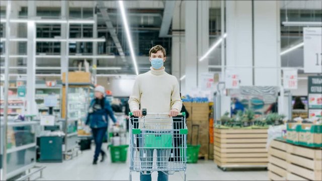 A man in a medical mask stands with a grocery cart in a supermarket, timelapse. Protection from coronavirus, purchase of essential goods.