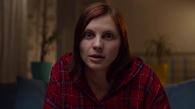 Young 30s woman in red shirt talking to camera sitting on sofa at home. Female telling stories looking at camera in slow motion. 