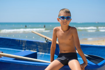A young boy in a blue boat on the ocean. A child in sunglasses on the beach near the shore.