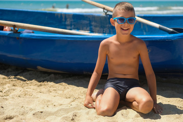 A young boy in a blue boat on the ocean. A child in sunglasses on the beach near the shore. © alexeytsurkan