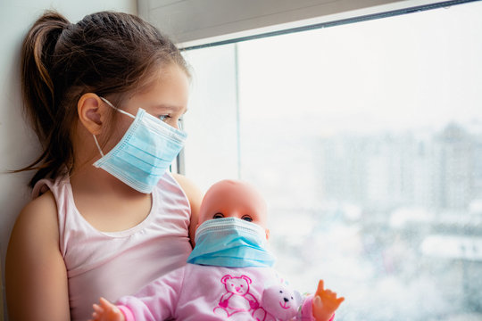 Little girl with doll wearing a protective mask. Copy space. Sadness kid at home in isolation. Sick child Little girls look at the window with longing on the street           