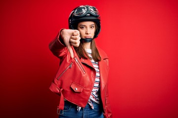 Young beautiful brunette motocyclist woman wearing motorcycle helmet and red jacket looking unhappy and angry showing rejection and negative with thumbs down gesture. Bad expression.