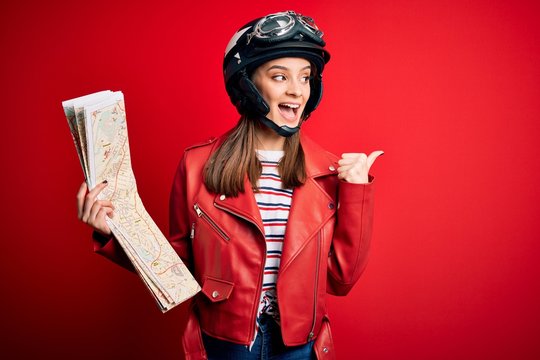 Young beautiful tourist motorcyclist woman wearing motorcycle helmet holding city map pointing and showing with thumb up to the side with happy face smiling
