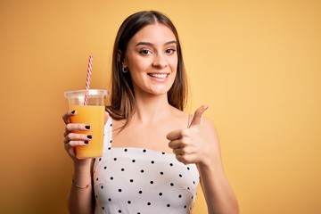 Young beautiful brunette woman drinking healthy orange juice over yellow background happy with big smile doing ok sign, thumb up with fingers, excellent sign