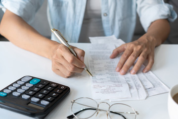 Pretty young Asian woman using a pen wriiting on the bills to calculate home expenses and taxes in...