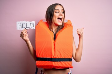 Young beautiful brunette woman wearing lifejacket holding paper with help word message pointing and...