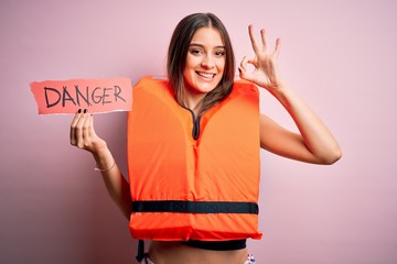 Young beautiful brunette woman wearing lifejacket holding paper with danger message doing ok sign...