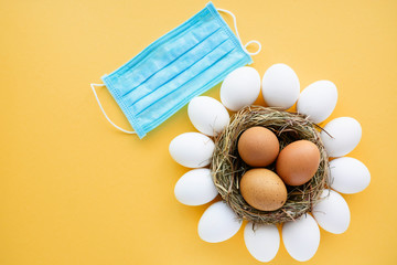 Easter colored eggs in the nest on yellow background with protective face mask - concept of...